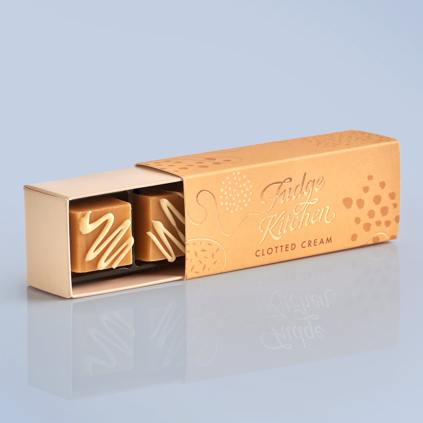 Gold fudge packaging with 'Fudge Kitchen' branding, slightly open to reveal a piece of clotted fudge a light blue background.