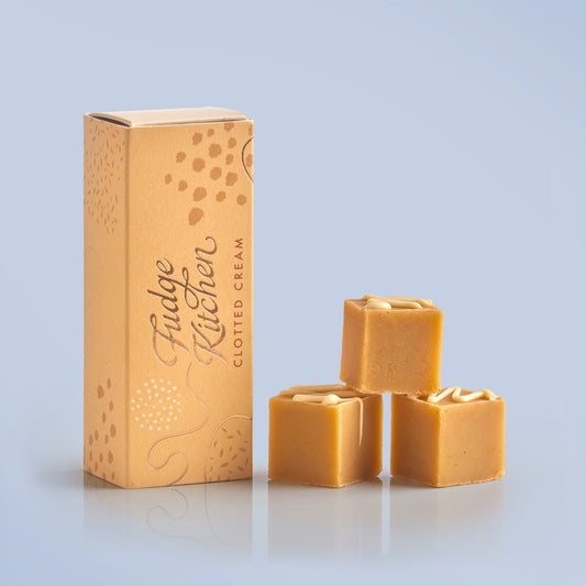 Three pieces of clotted cream fudges stacked in front of the long Fudge Kitchen yellow gift box on a pale blue background.
