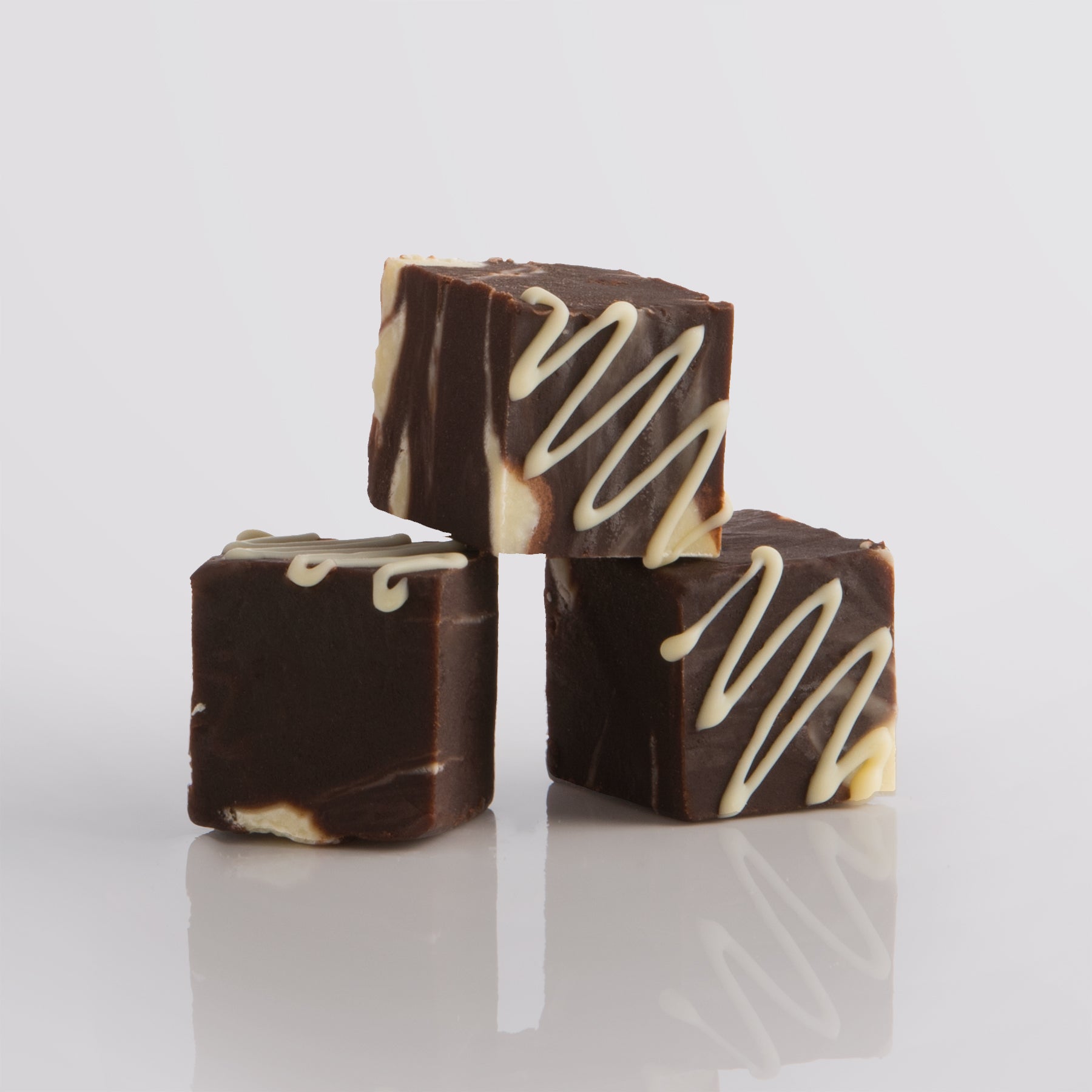 Three pieces of double chocolate fudge with white chocolate drizzle on a reflective surface.