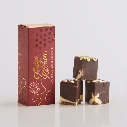 Three pieces of double chocolale fudges stacked in front of the long Fudge Kitchen  gift box on a grey background.