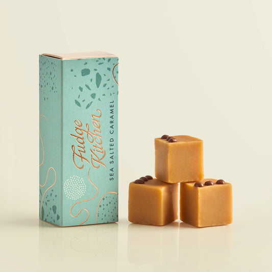 Three pieces of salted caramel fudges stacked in front of the long Fudge Kitchen gift box on a beige background.