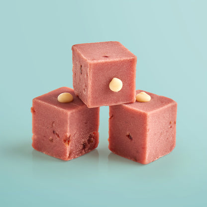 Three pink strawberries and cream fudge cubes with white chocoalte drops on a light blue background.