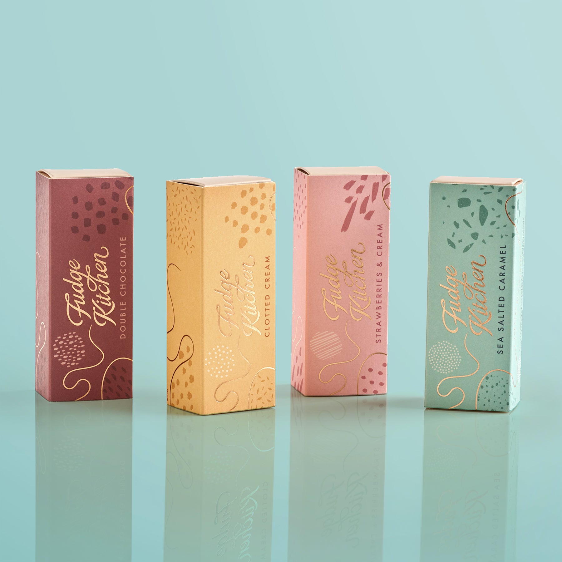 Four colourful fudge slim slider boxes of Fudge Kitchen fudge selections on a light blue background.