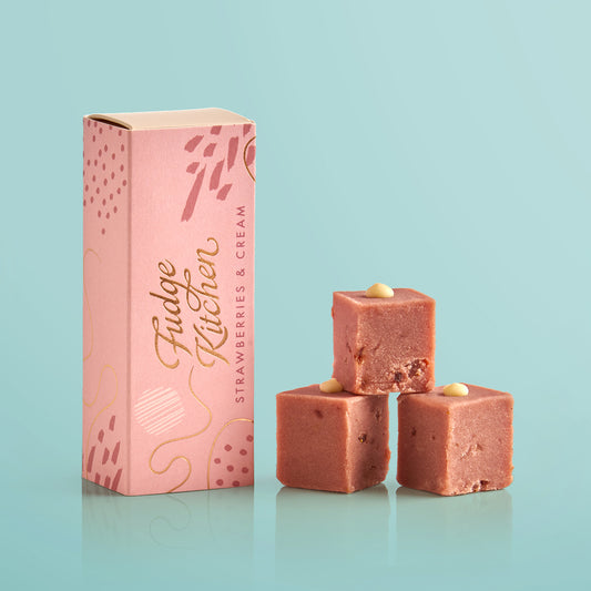 Three pieces of strawberries & cream fudges stacked in front of the long Fudge Kitchen christmas gift box on a blue background.
