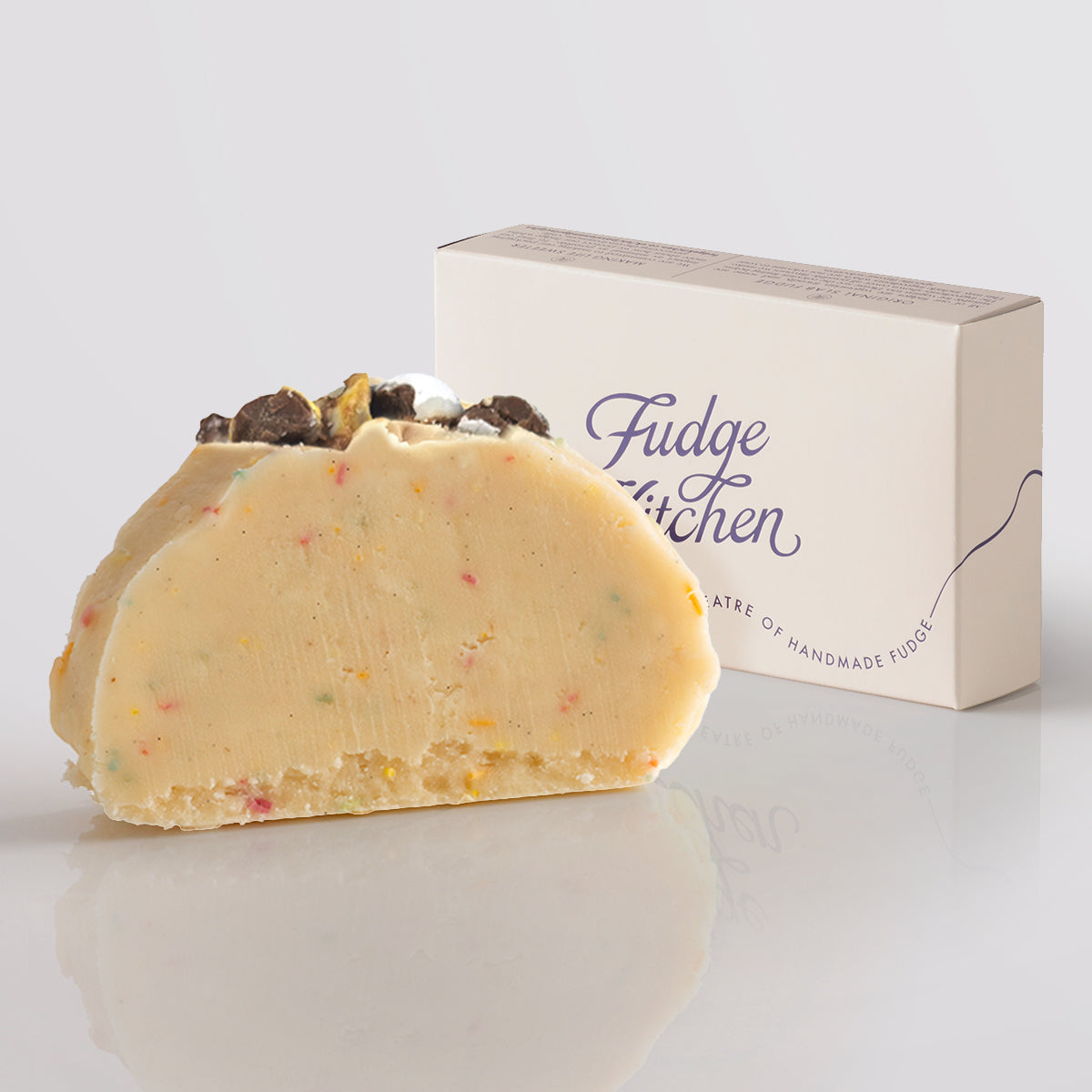 A slice of Speckled Egg Fudge with colourful sprinkles, topped with crushed chocolate eggs, next to a 'Fudge Kitchen' box on a light gray background.