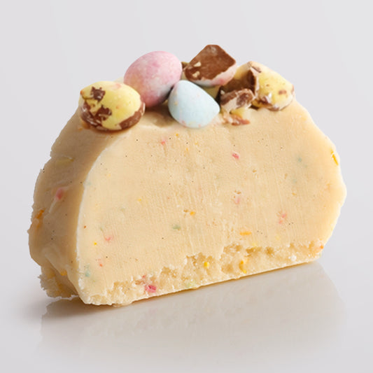 A slice of Speckled Egg Fudge with colourful candy pieces and topped with crushed chocolate eggs, on a light gray background.