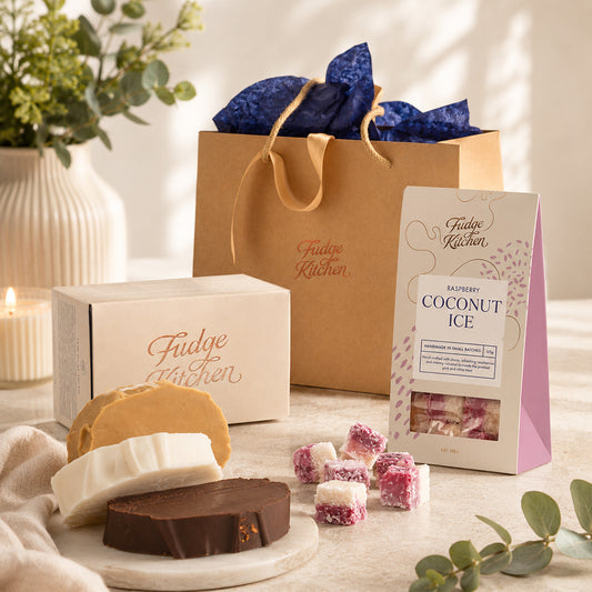 Fudge Kitchen vegan sweet gifts and confectionery, including a box, 3 slices of fudge, gift bag, and raspberry coconut ice on a table with a plant in the background.
