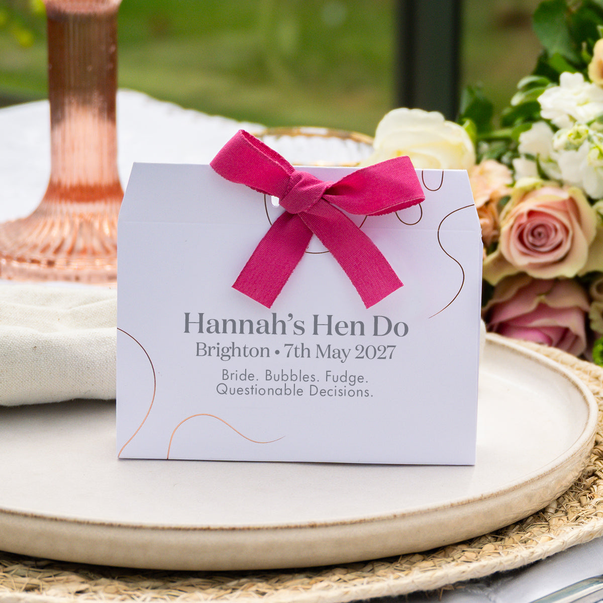 A white bronze foil embellished favour tied with a bright pink ribbon sits on a woven tray in an outdoor setting with flowers and a copper-toned glass in the background. The card reads: 'Hannah’s Hen Do, Brighton • 7th May 2027, Bride, Bubbles, Fudge, Questionable Decisions.