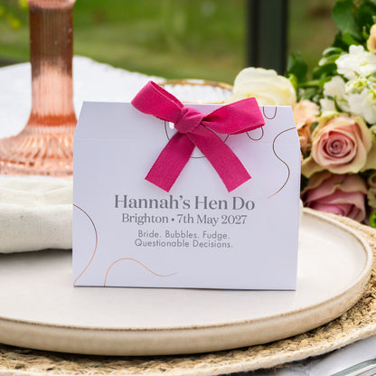 A white bronze foil embellished favour tied with a bright pink ribbon sits on a woven tray in an outdoor setting with flowers and a copper-toned glass in the background. The card reads: 'Hannah’s Hen Do, Brighton • 7th May 2027, Bride, Bubbles, Fudge, Questionable Decisions.