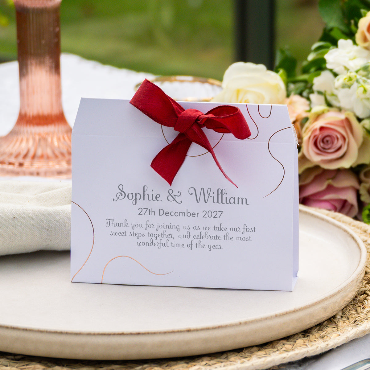 A personalised wedding favour box with a white card, elegant copper line art, and a red ribbon bow, displayed on a woven placemat with flowers and a glass in the background.