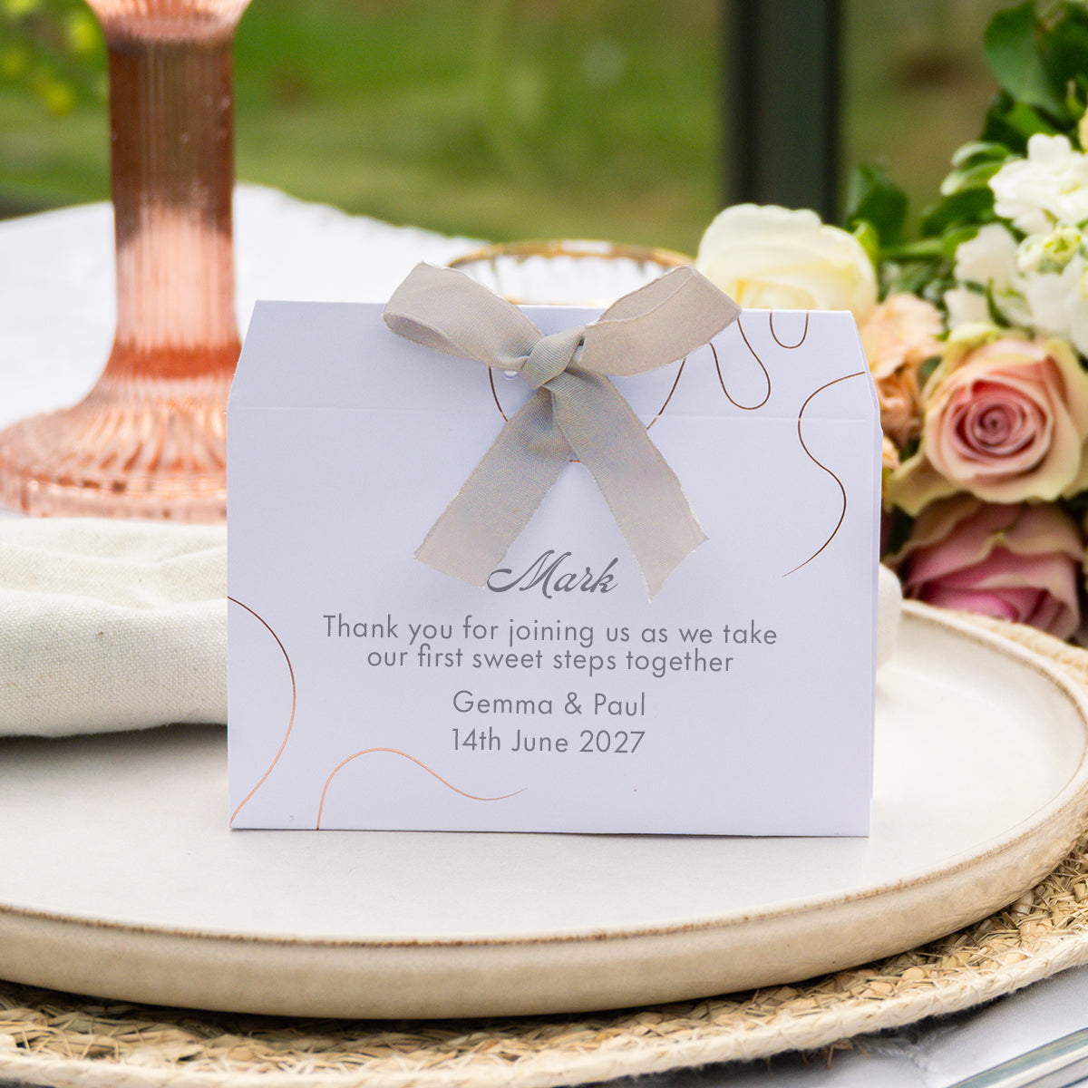 A personalised wedding favour box with  elegant rose gold line art, and a light beige satin ribbon bow. The front is printed with a custom thank you message for 'Mark' dated 14th June 2027.