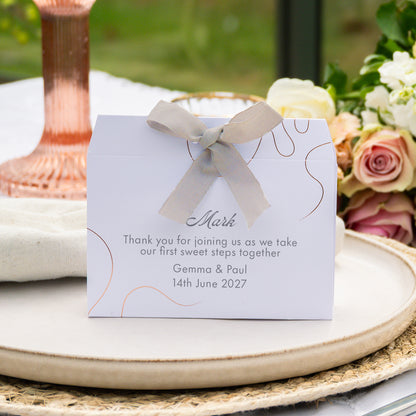 A personalised wedding favour box with  elegant rose gold line art, and a light beige satin ribbon bow. The front is printed with a custom thank you message for 'Mark' dated 14th June 2027.