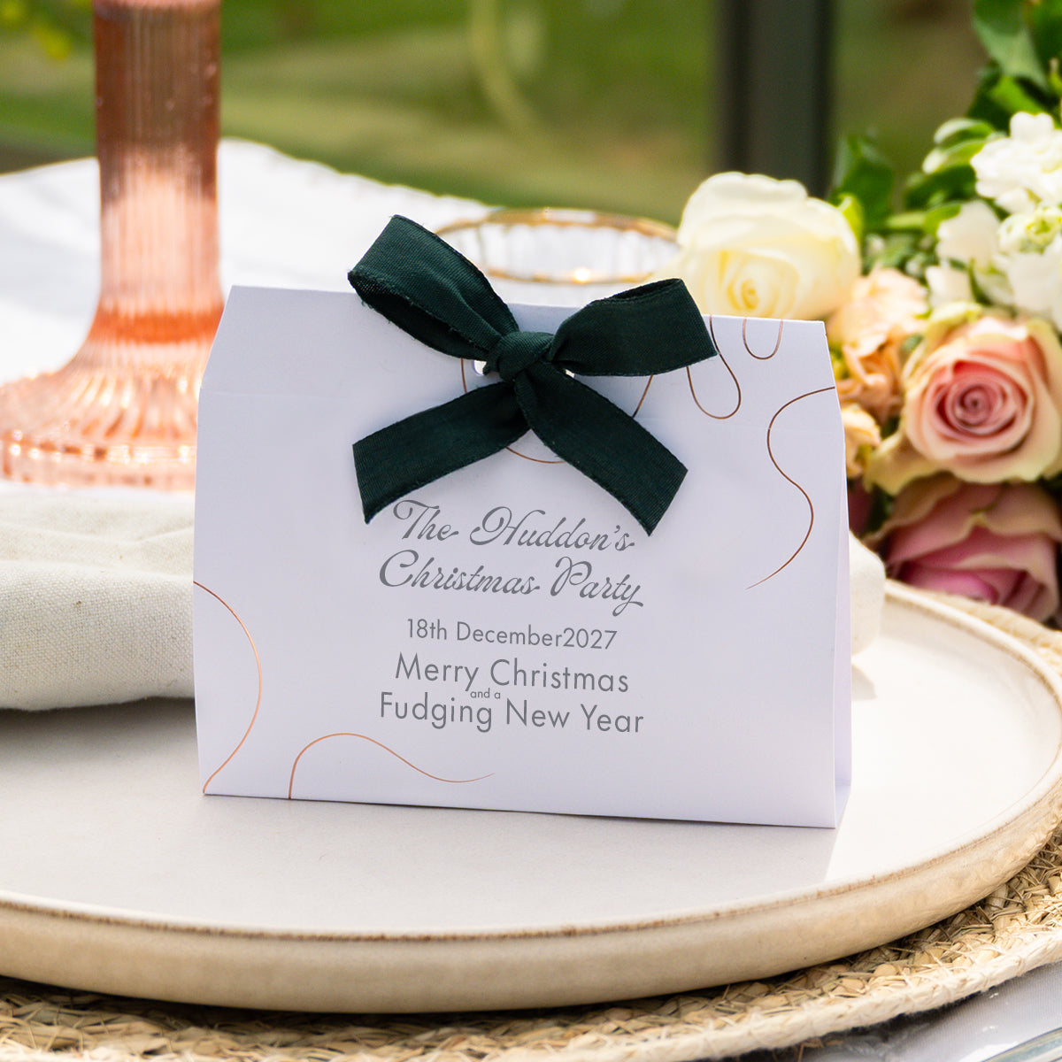 A personalised Christmas party favor box with a dark green satin bow, placed on a styled table setting with flowers and a copper-toned candlestick.