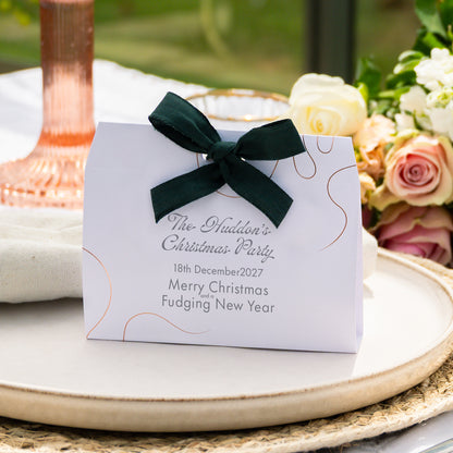 A personalised Christmas party favor box with a dark green satin bow, placed on a styled table setting with flowers and a copper-toned candlestick.