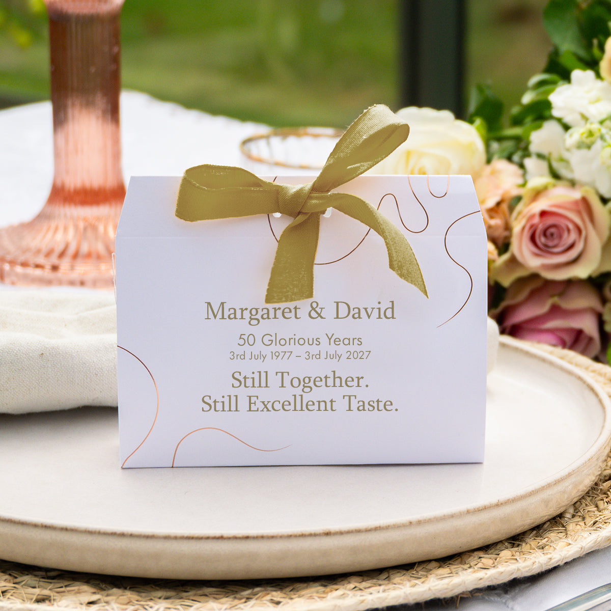 A personalised white wedding favour box tied with a gold ribbon, placed on a ceramic tray with flowers and tableware in the background.
