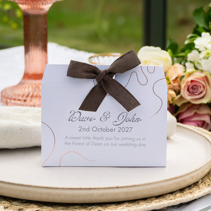 A personalised wedding favour box with a dark brown ribbon bow, featuring elegant script text that reads 'Dave & John 2nd October 2027' and a sweet thank you message. The box is styled on a beige ceramic plate with a blurred rose arrangement and a copper-colored candlestick in the background.