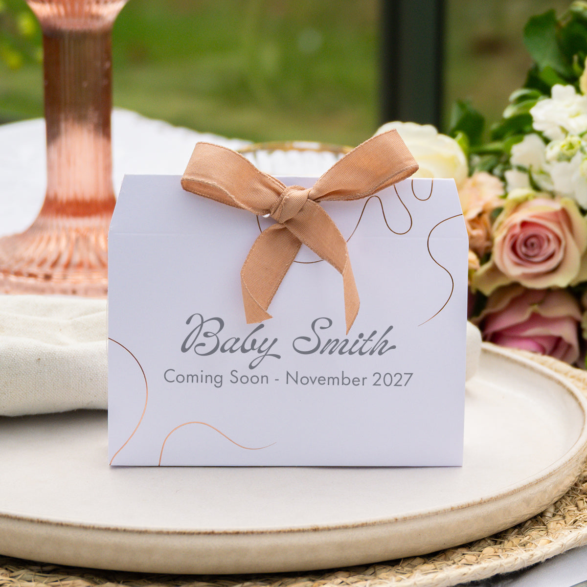 A personalised baby shower favour box with a mink ribbon bow, printed with 'Baby Smith' and 'Coming Soon - November 2027' on a white background with floral and table setting props.