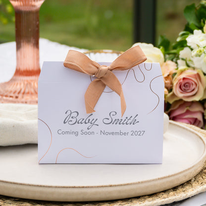 A personalised baby shower favour box with a mink ribbon bow, printed with 'Baby Smith' and 'Coming Soon - November 2027' on a white background with floral and table setting props.