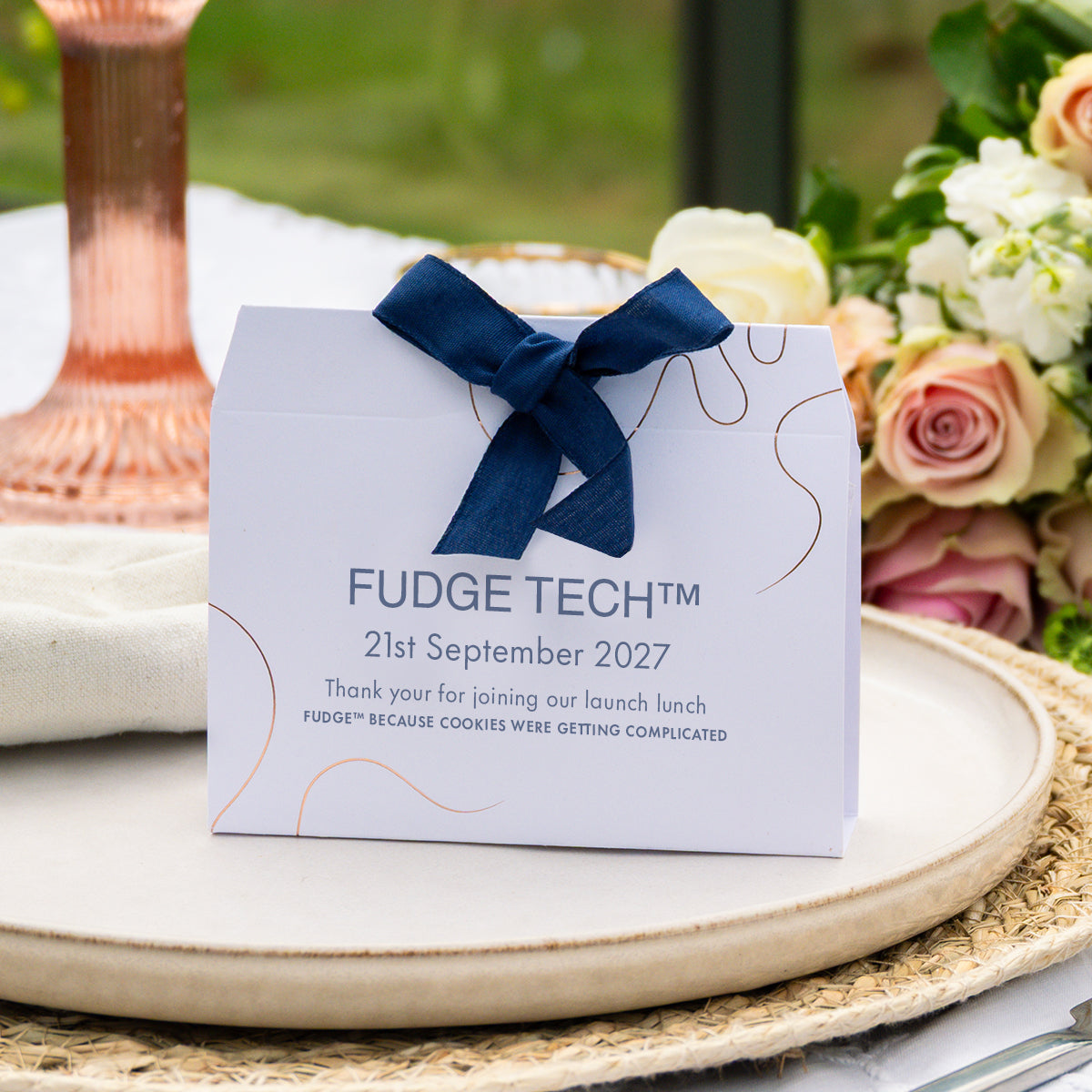 A white Fudge Fresh thank-you event favour with a navy blue ribbon bow, styled on a woven placemat with flowers and a copper candlestick in the background.