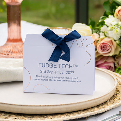 A white Fudge Fresh thank-you event favour with a navy blue ribbon bow, styled on a woven placemat with flowers and a copper candlestick in the background.