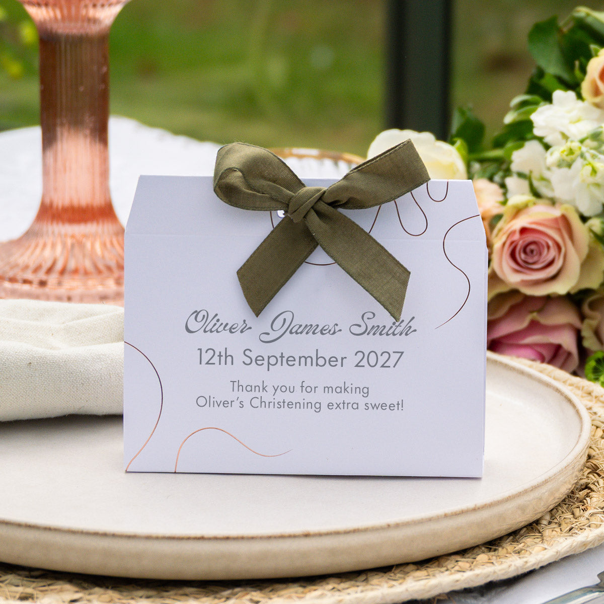 A personalised favour box card with an olive green ribbon bow, placed on a styled wedding table setting with flowers and a candle.