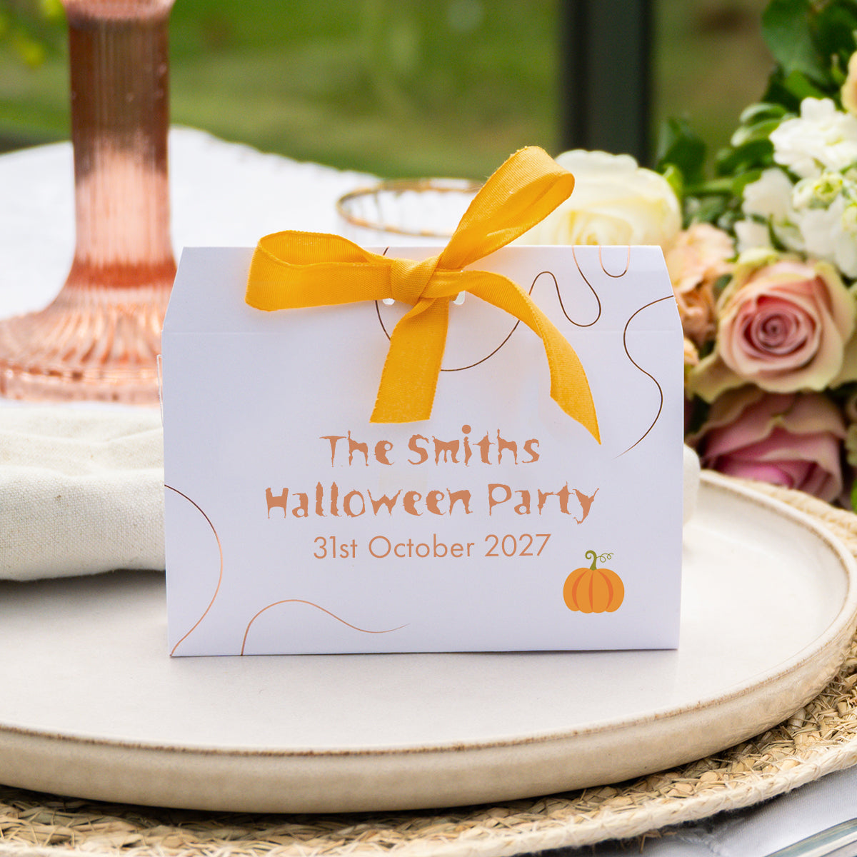 A white favour box tied with a bright yellow ribbon sits on a round beige tray. The front of the box is printed with the text 'The Smiths Halloween Party 31st October 2027' in orange, along with thin bronze decorative embellishment and a small orange pumpkin illustration.