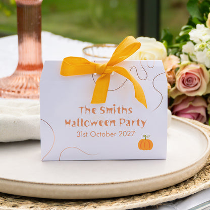 A white favour box tied with a bright yellow ribbon sits on a round beige tray. The front of the box is printed with the text 'The Smiths Halloween Party 31st October 2027' in orange, along with thin bronze decorative embellishment and a small orange pumpkin illustration.