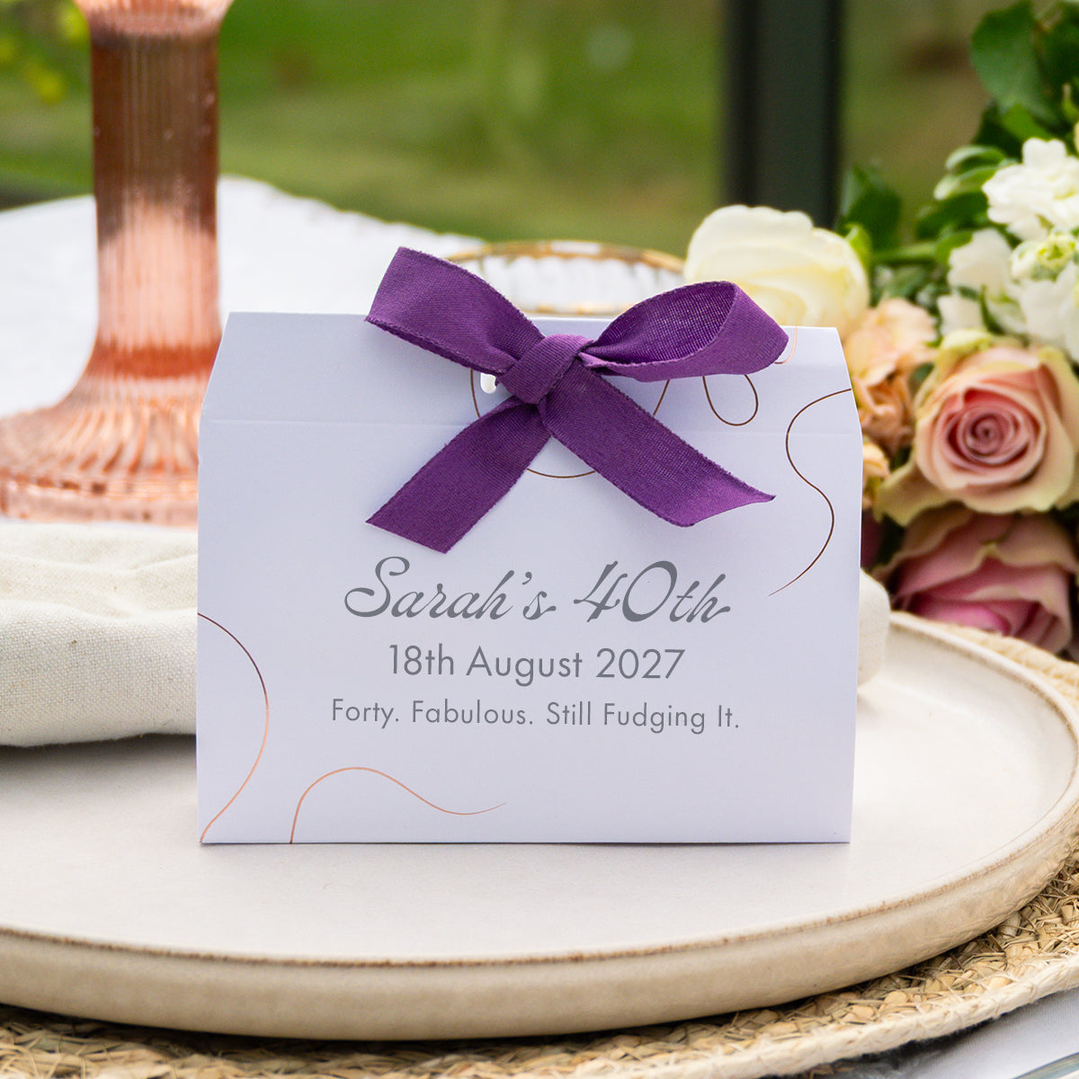 A white wedding favour box tied with a purple ribbon, printed with the text 'Sarah's 40th 18th August 2027 Forty. Fabulous.