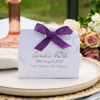 A white wedding favour box tied with a purple ribbon, printed with the text 'Sarah's 40th 18th August 2027 Forty. Fabulous.