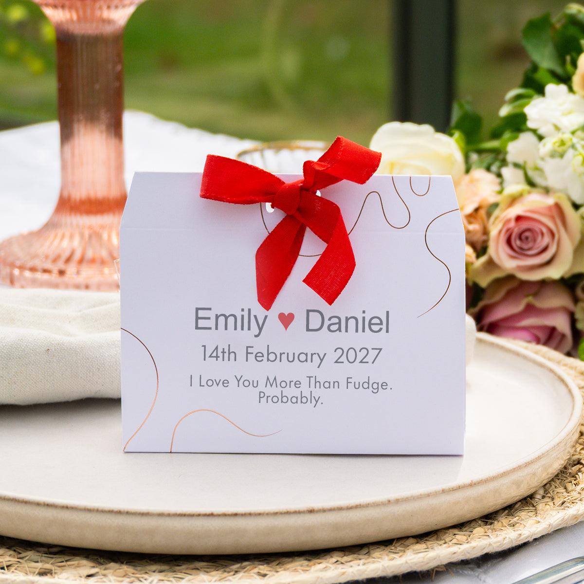A personalised white wedding favour tied with a red ribbon, displayed on a beige tray with flowers and a rose gold candle in the background.