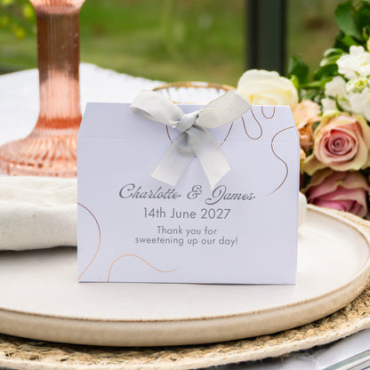 A personalised wedding favour tied with a white satin ribbon, displayed on a ceramic tray with a rose gold candlestick and floral arrangement in the background.