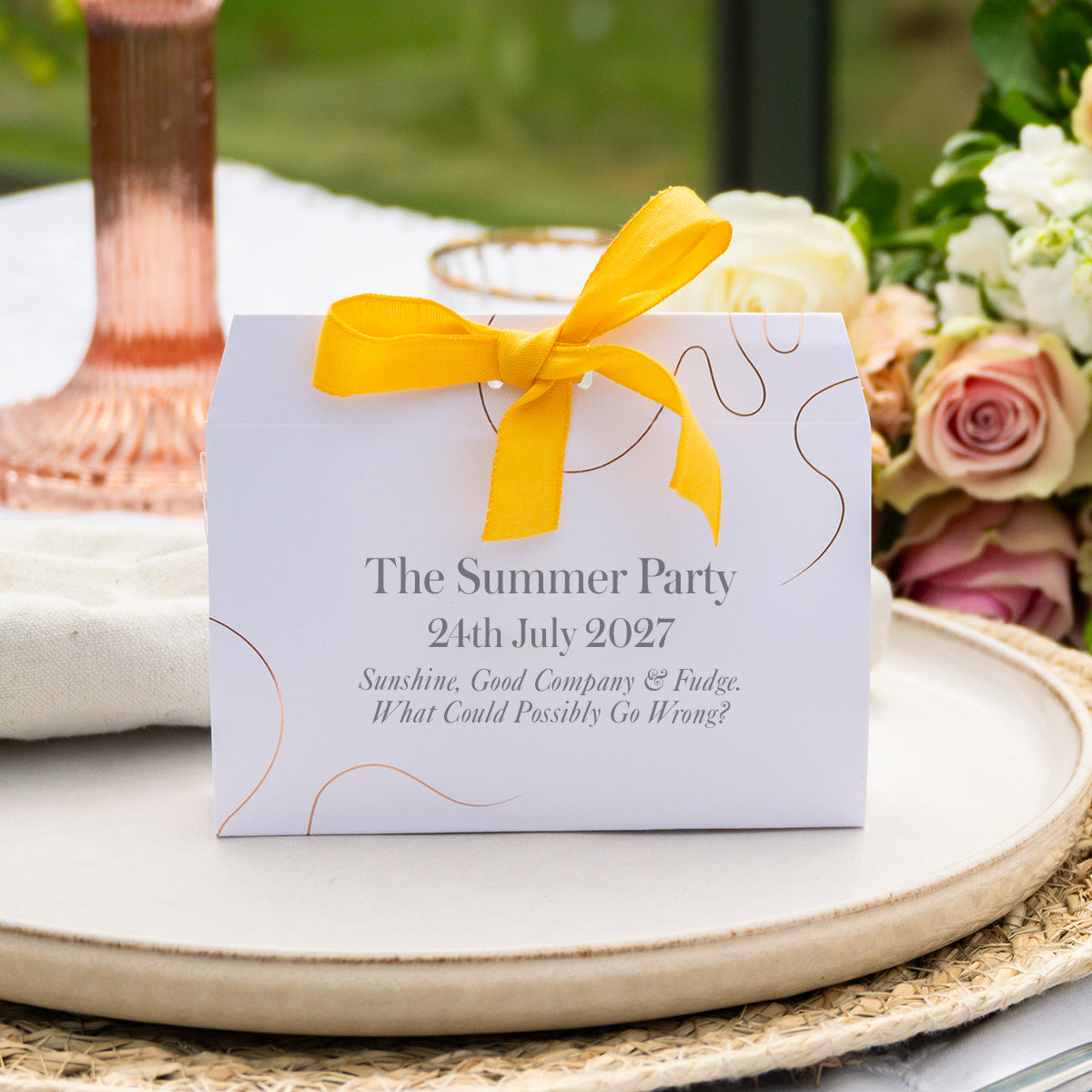 A white wedding favour box tied with a bright yellow ribbon, printed with the text 'The Summer Party 24th July 2027' and a small gold abstract line design, styled on a ceramic tray with flowers in the background.