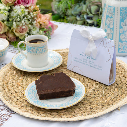 A personalized favour with a white satin ribbon sits on a woven placemat next to a plate of chocolate slice of fudge and a matching coffee cup on a table with flowers in the background.