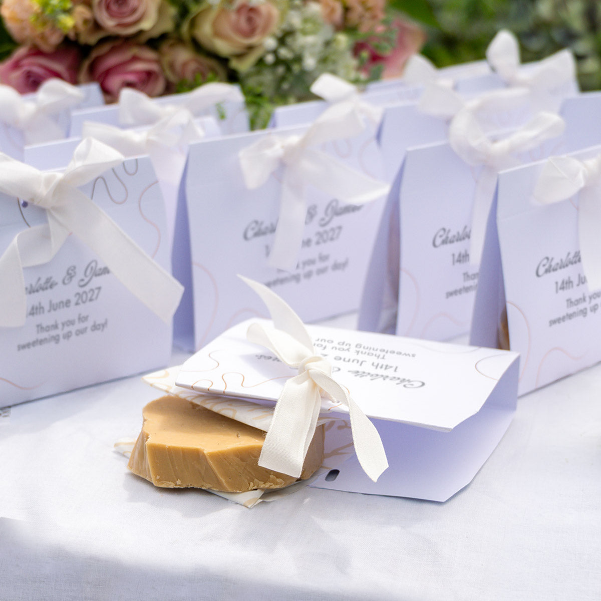 Personalised fudge favours tied with white ribbons, arranged on a white tablecloth with a slice of fudge in the front. The bags are printed with names and date, with blurred flowers in the background.