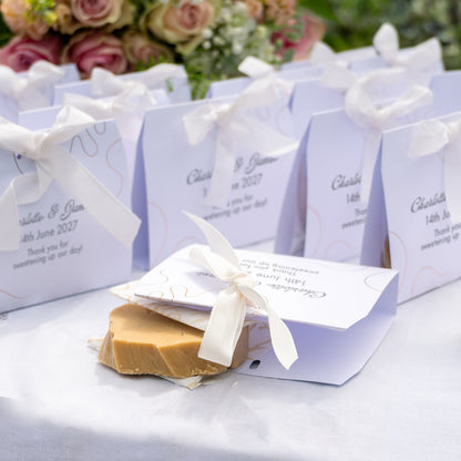 Personalised fudge favours tied with white ribbons, arranged on a white tablecloth with a slice of fudge in the front. The bags are printed with names and date, with blurred flowers in the background.