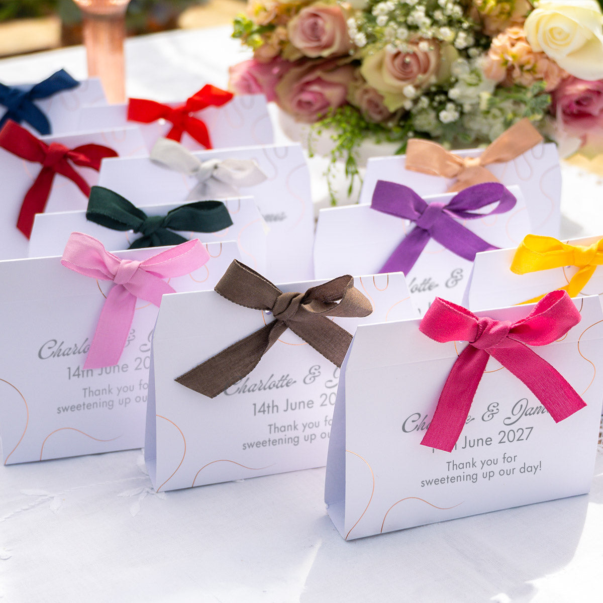 A set of personalised white favour bags tied with colorful satin ribbons in pink, brown, purple, red, blue, green, and yellow, arranged on a white tablecloth with a blurred floral arrangement in the background.