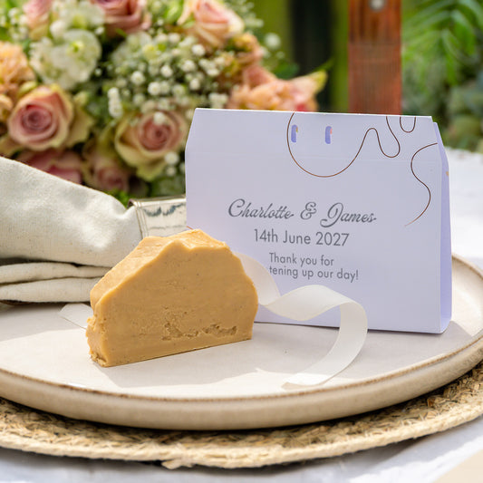 A custom wedding favour envelope with a slice of fudge, displayed on a ceramic plate with a matching paper tag that reads 'Charlotte & James 14th June 2027'.