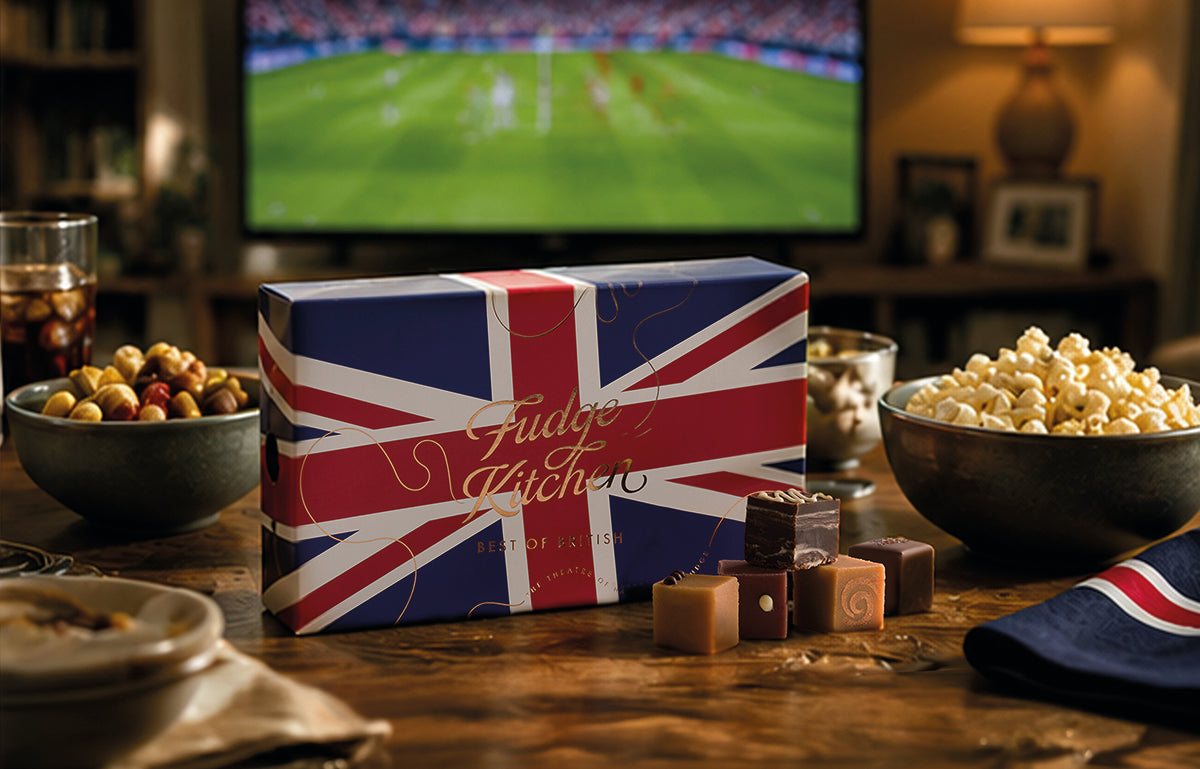 Fudge Kitchen fudge selection box with Union Jack design on a table with popcorn and snacks, TV in the background.