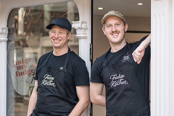 Two men wearing black aprons with 'Fudge Kitchen' written in white script, standing in a doorway. The man on the left wears a black cap and smiles, while the man on the right wears a tan cap.